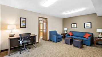 A hotel room with a desk, office chair, blue sofa, two side tables with lamps, and a carpeted floor.