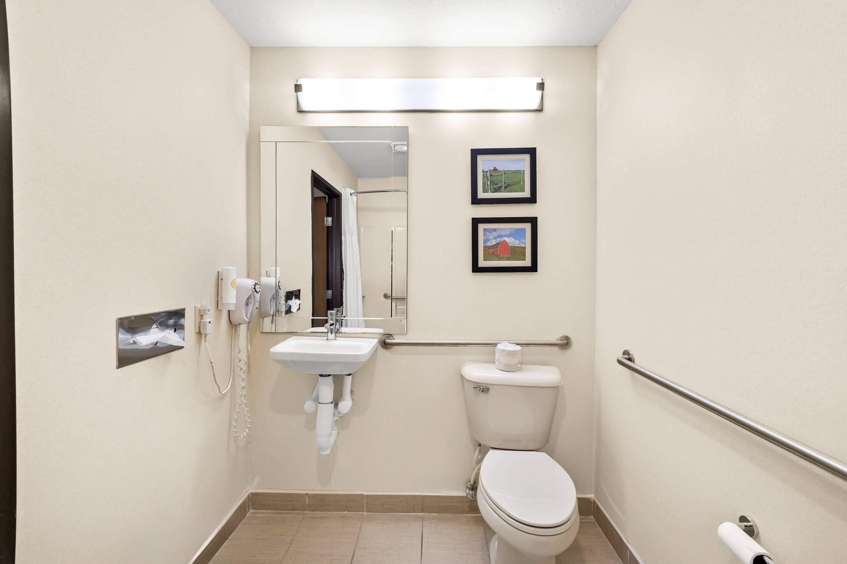 Un baño pequeño y bien iluminado con lavamanos, inodoro y secador de manos. Hay dos cuadros en la pared y un espejo sobre el lavamanos.