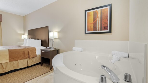A hotel room with a large white bathtub, a bed with a brown blanket, a nightstand with a lamp, and a framed picture on the wall. - Best Western Teal Lake Inn (Mexico)