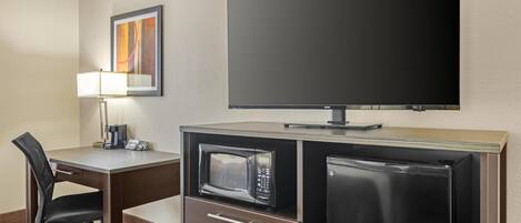Een compacte hotelkamer met een bureau, stoel, magnetron, koelkast en een wandgemonteerde flatscreen-tv.
