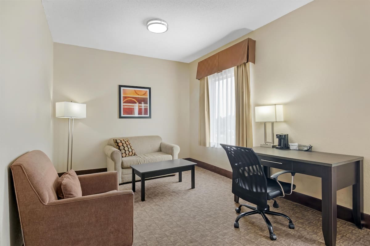 A hotel room with a sofa, a desk, a chair, a coffee table, a lamp, and a painting on the wall.