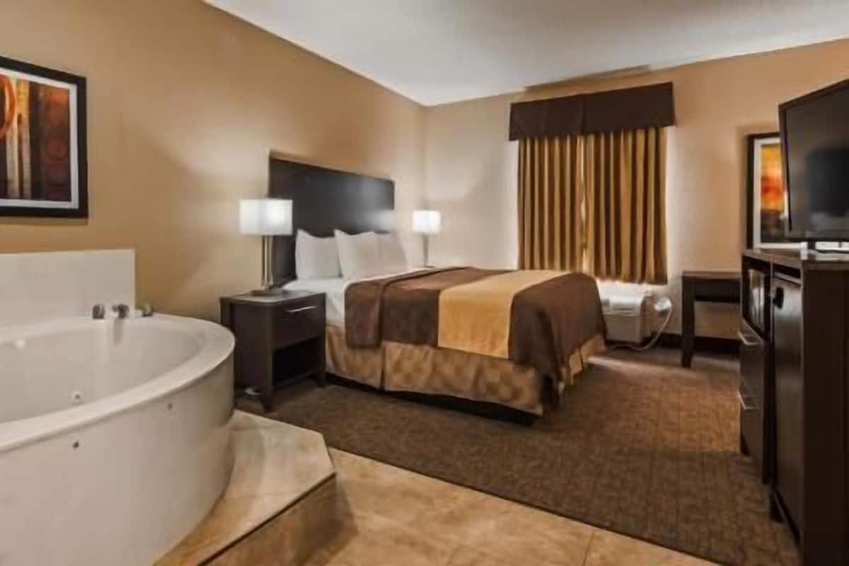A hotel room with a large bathtub, a bed with a brown blanket, a television, and a painting on the wall.