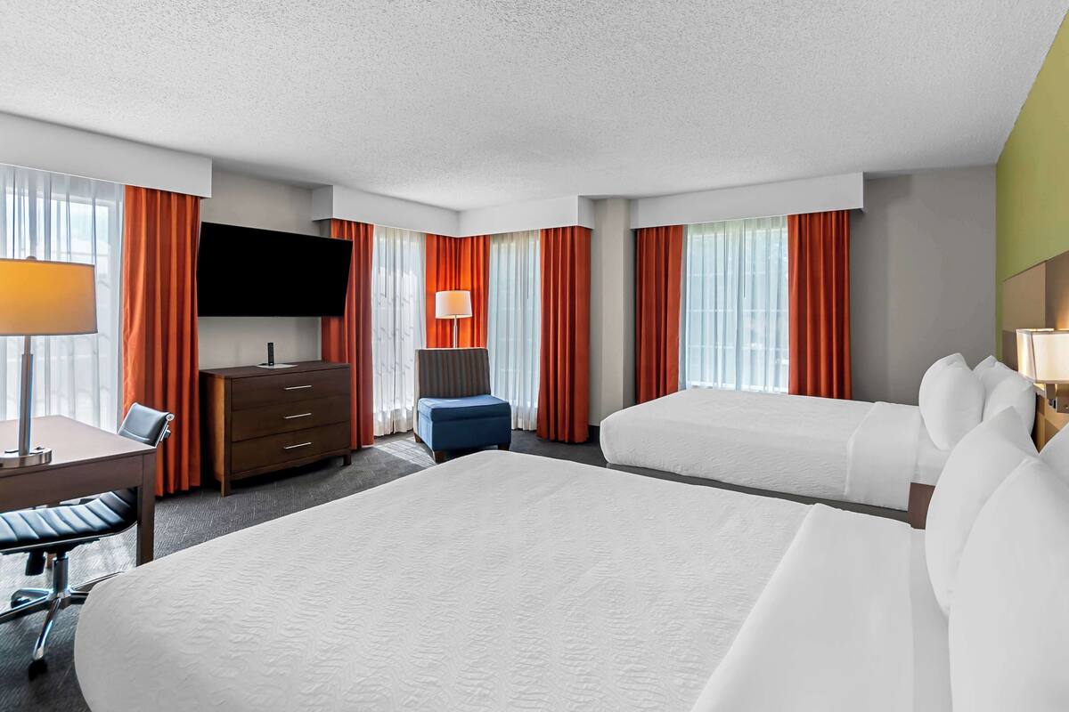 Ein Hotelzimmer mit zwei Betten, einem Schreibtisch mit Stuhl, einem an der Wand befestigten Fernseher und einem Fenster mit Vorhängen.