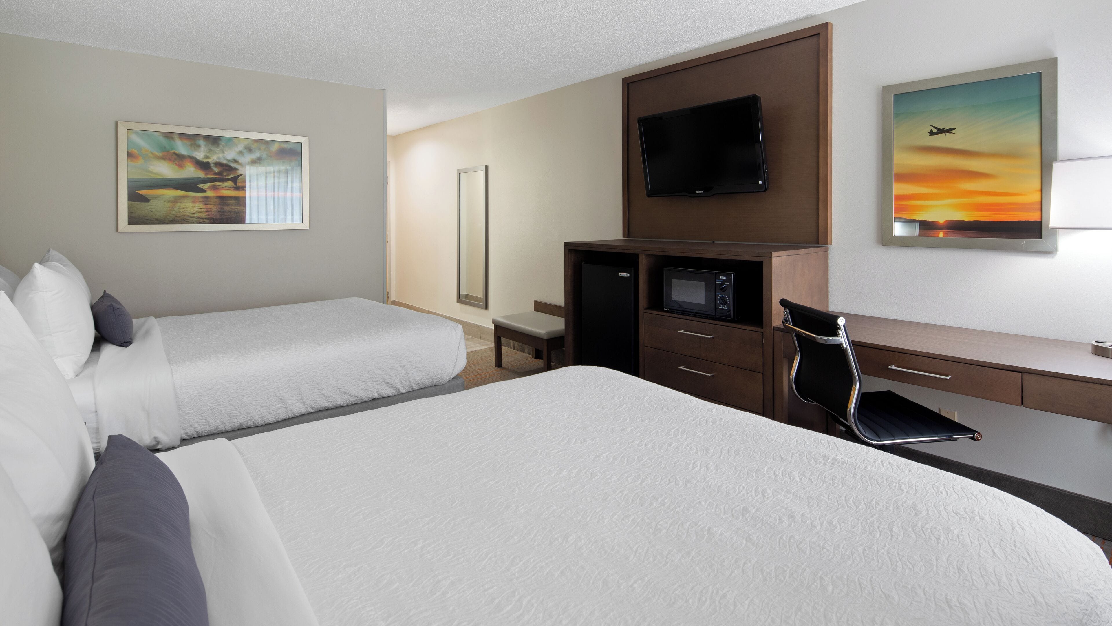 A hotel room with two beds, a desk, a chair, a TV, and artwork on the walls.