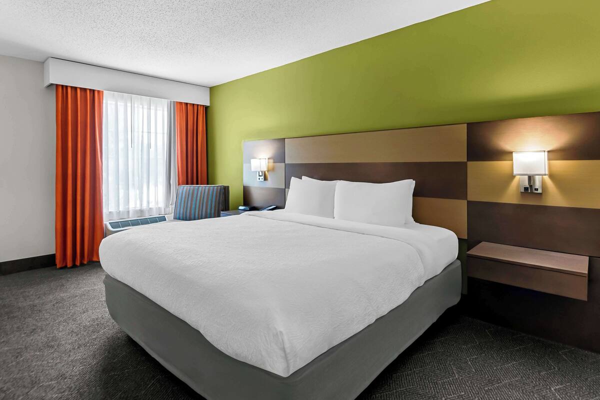 Ein Hotelzimmer mit einem großen Bett, einem Nachttisch, einer Lampe und einem Fenster mit Vorhängen.