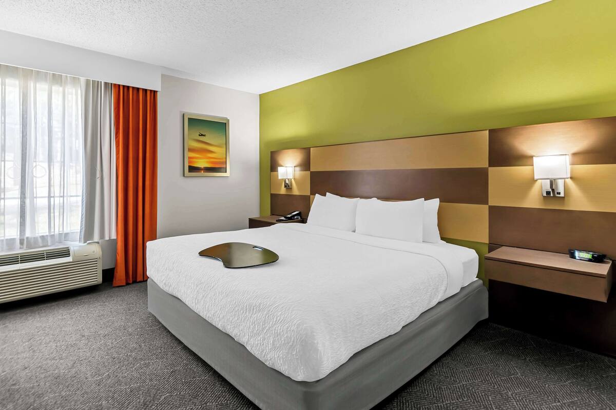 Ein Hotelzimmer mit einem großen Bett, einem Nachttisch mit Lampe, einer Wand-Klimaanlage und einem Fenster mit Vorhängen.
