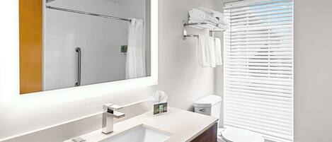 Un baño moderno con un espejo grande, lavamanos blanco y retrete. Se observa un perchero con toallas dobladas y una ventana con persianas.