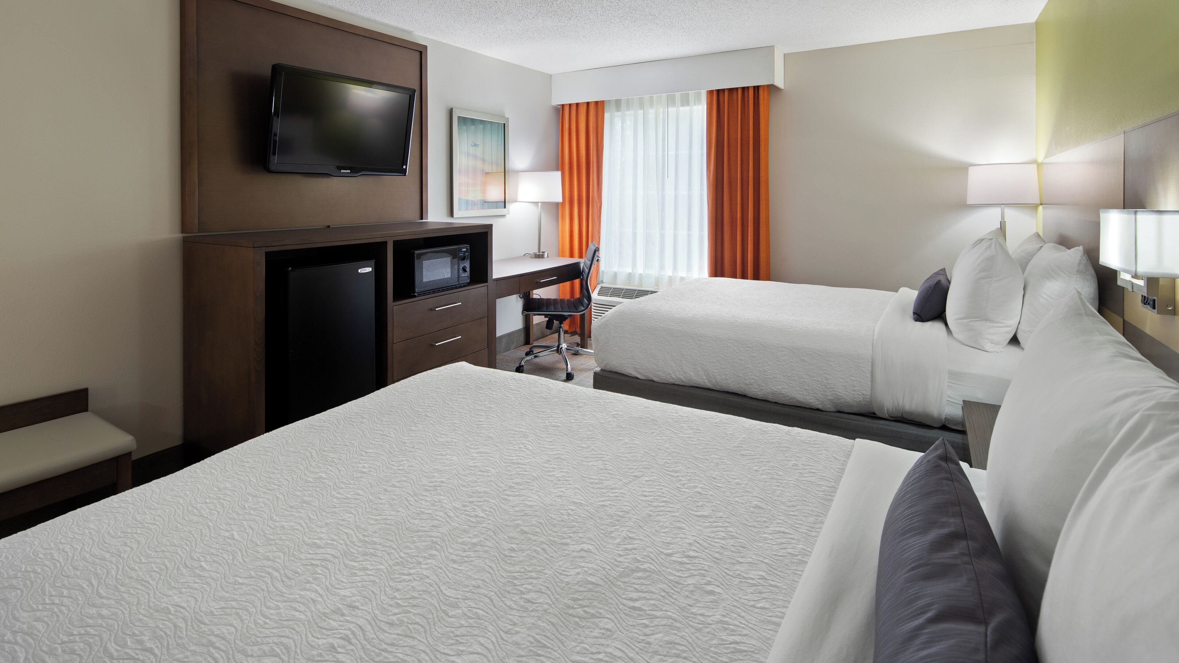 A hotel room with two beds, a desk with a television, a refrigerator, and a chair.