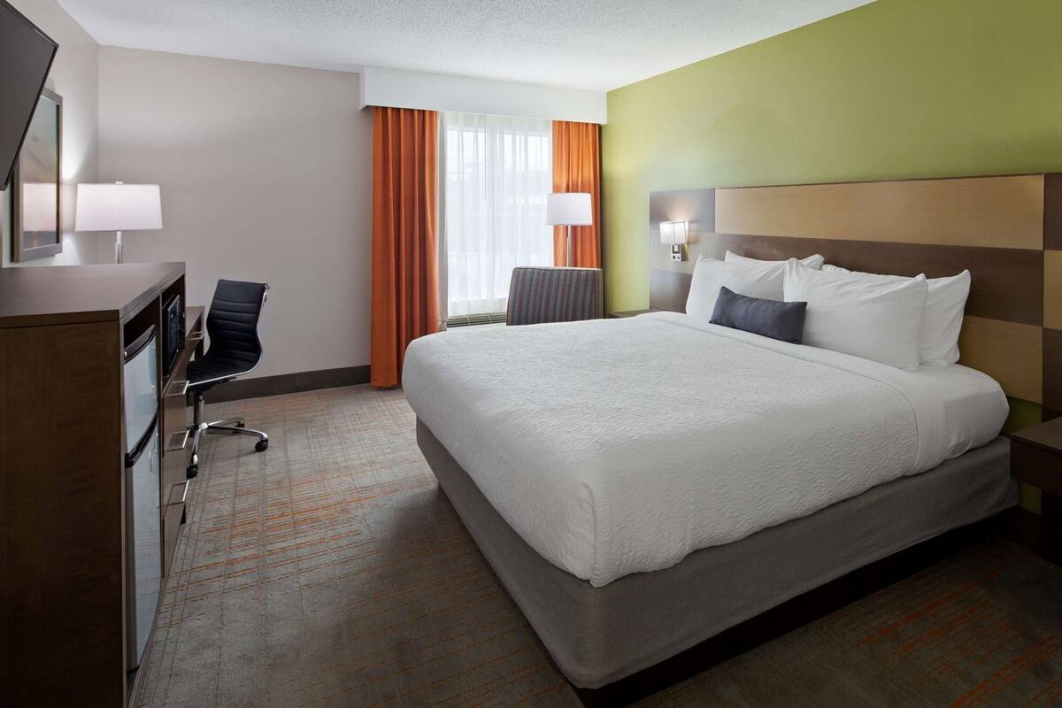 Ein Hotelzimmer mit einem großen Bett, einem Schreibtisch mit Stuhl, einer Lampe und einem Fenster mit Vorhängen.