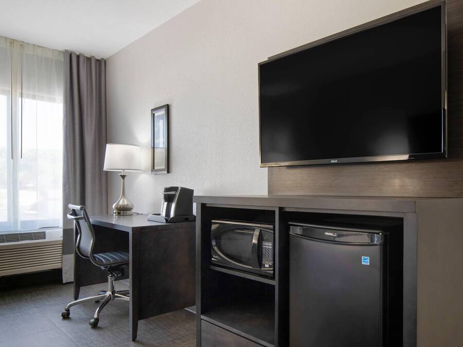 A hotel room with a flat-screen TV, a desk with a lamp, a chair, a microwave, and a coffee maker.