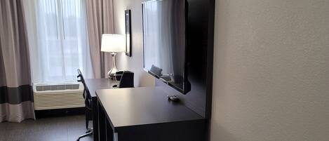 A hotel room with a flat-screen TV mounted on the wall, a desk with a chair, a lamp, and a window with curtains.