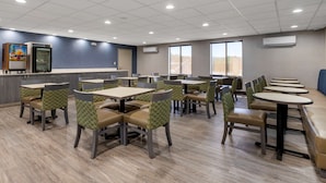 A modern dining area with multiple tables and chairs, a counter with a refrigerator, and large windows providing natural light.