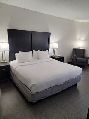 A neatly made bed with white linens, a dark headboard, two bedside lamps, a chair, and a nightstand with a phone.