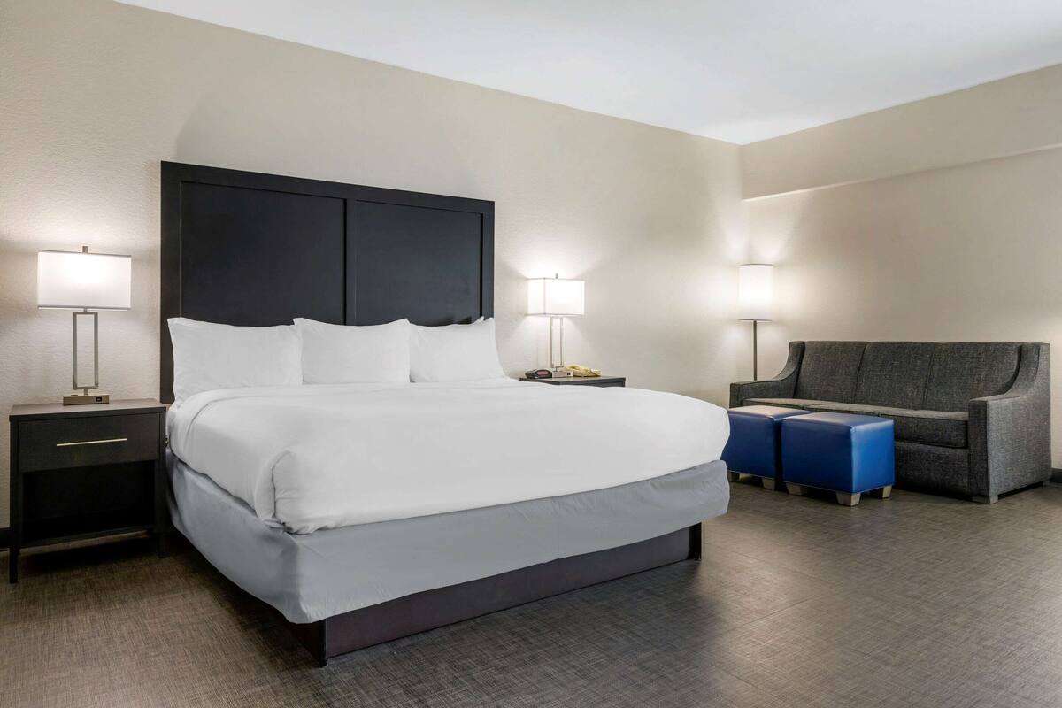A hotel room with a large bed, bedside lamps, a nightstand, a sofa, and two ottomans.