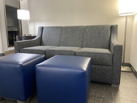 Un sofá tapizado en gris con un otoman correspondiente, un espejo y una lámpara en una habitación de hotel.