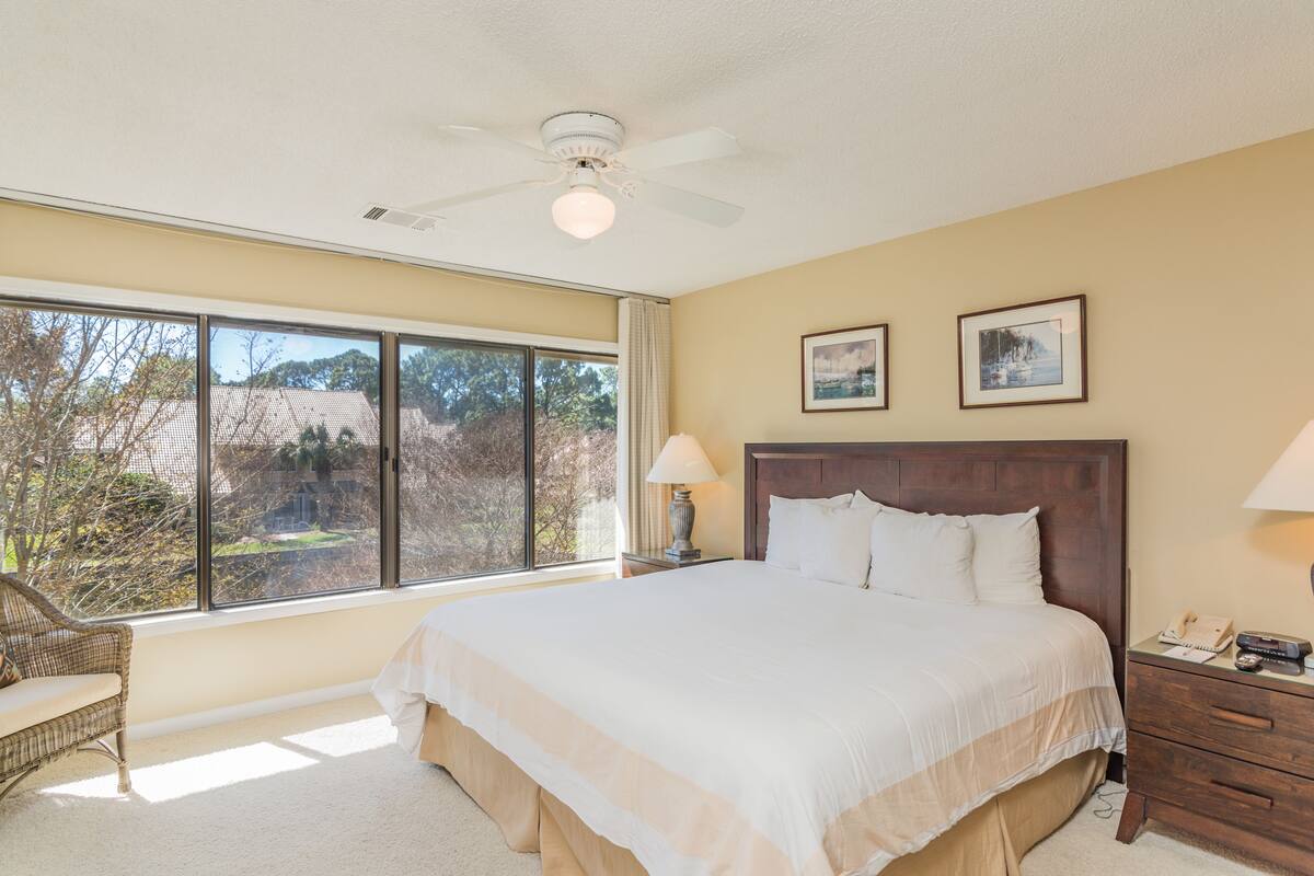 A bedroom with a large bed, a ceiling fan, a window with a view of trees, and a nightstand with a lamp.