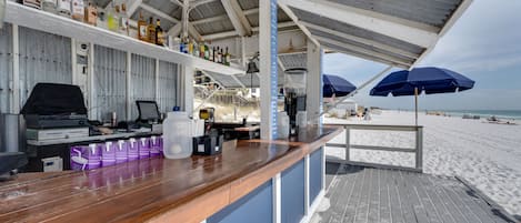 A beachside bar with wooden counters, a grill, and a view of the beach.
