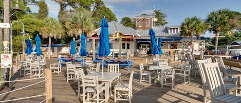 A wooden deck with tables and chairs, blue umbrellas, and a building with a blue roof in the background.