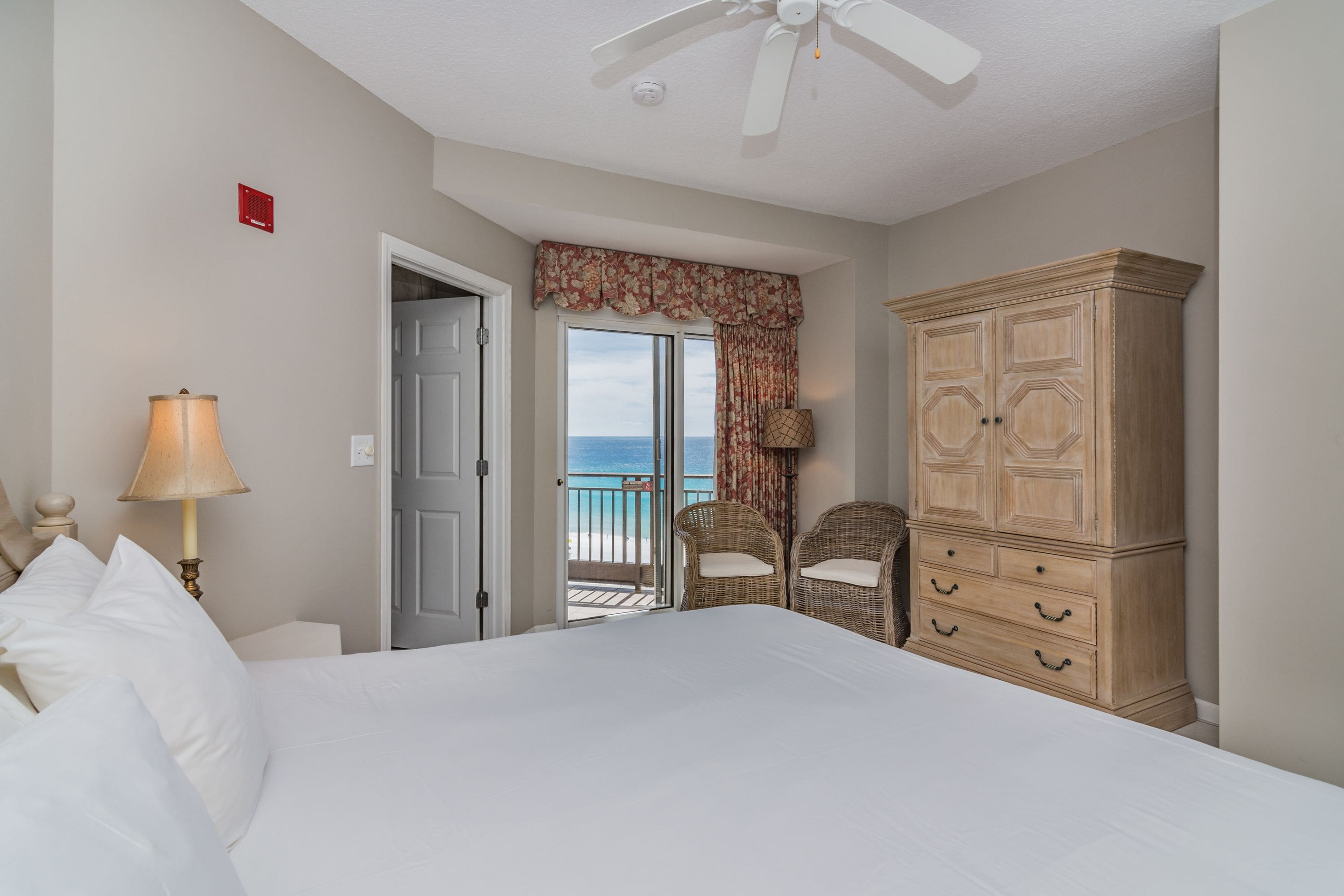 Une chambre avec un grand lit, une armoire, deux fauteuils en osier, une lampe et une vue sur la mer par une porte.