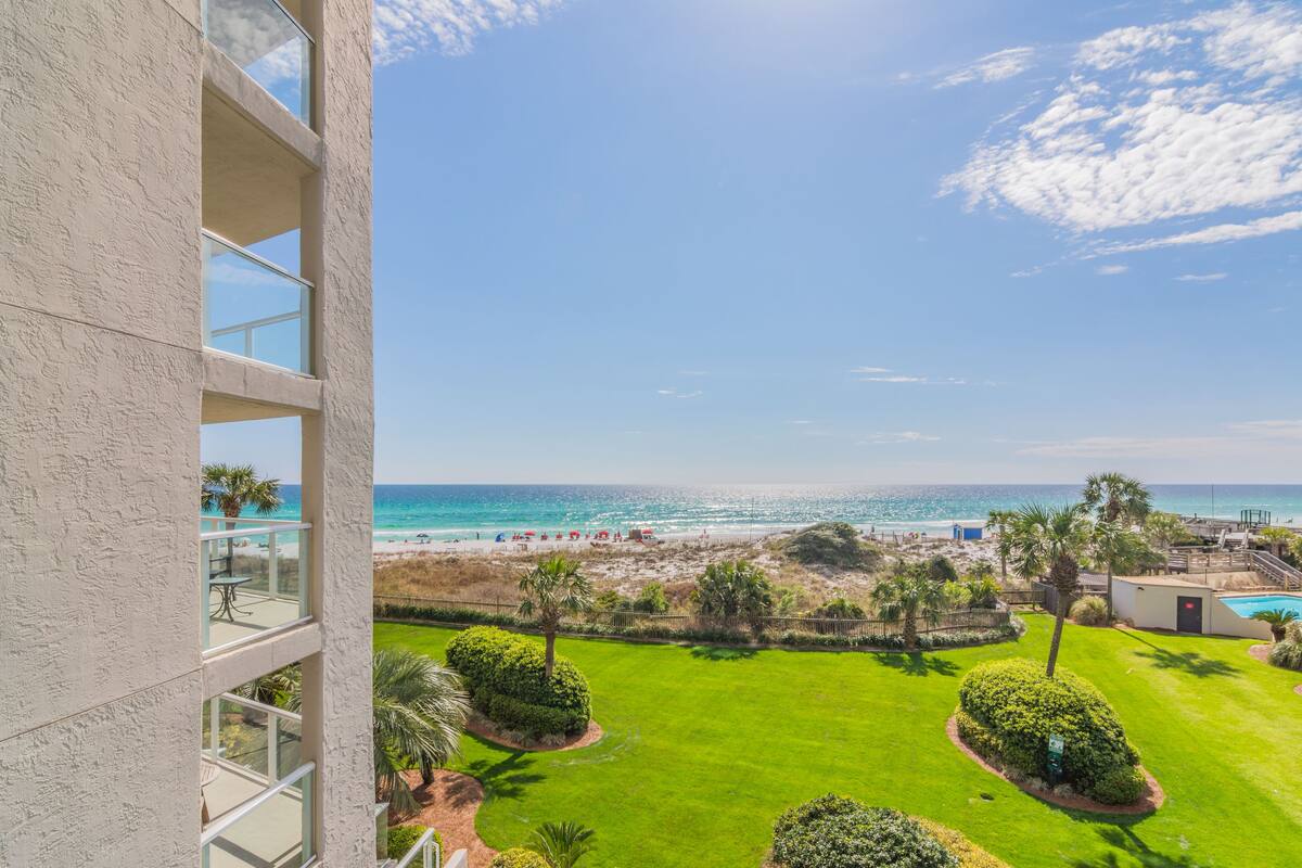 A view from a balcony overlooking a beach with a clear sky, a pool, and well-maintained lawns.
