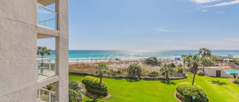 A view from a balcony overlooking a beach with a clear sky, a pool, and well-maintained lawns.