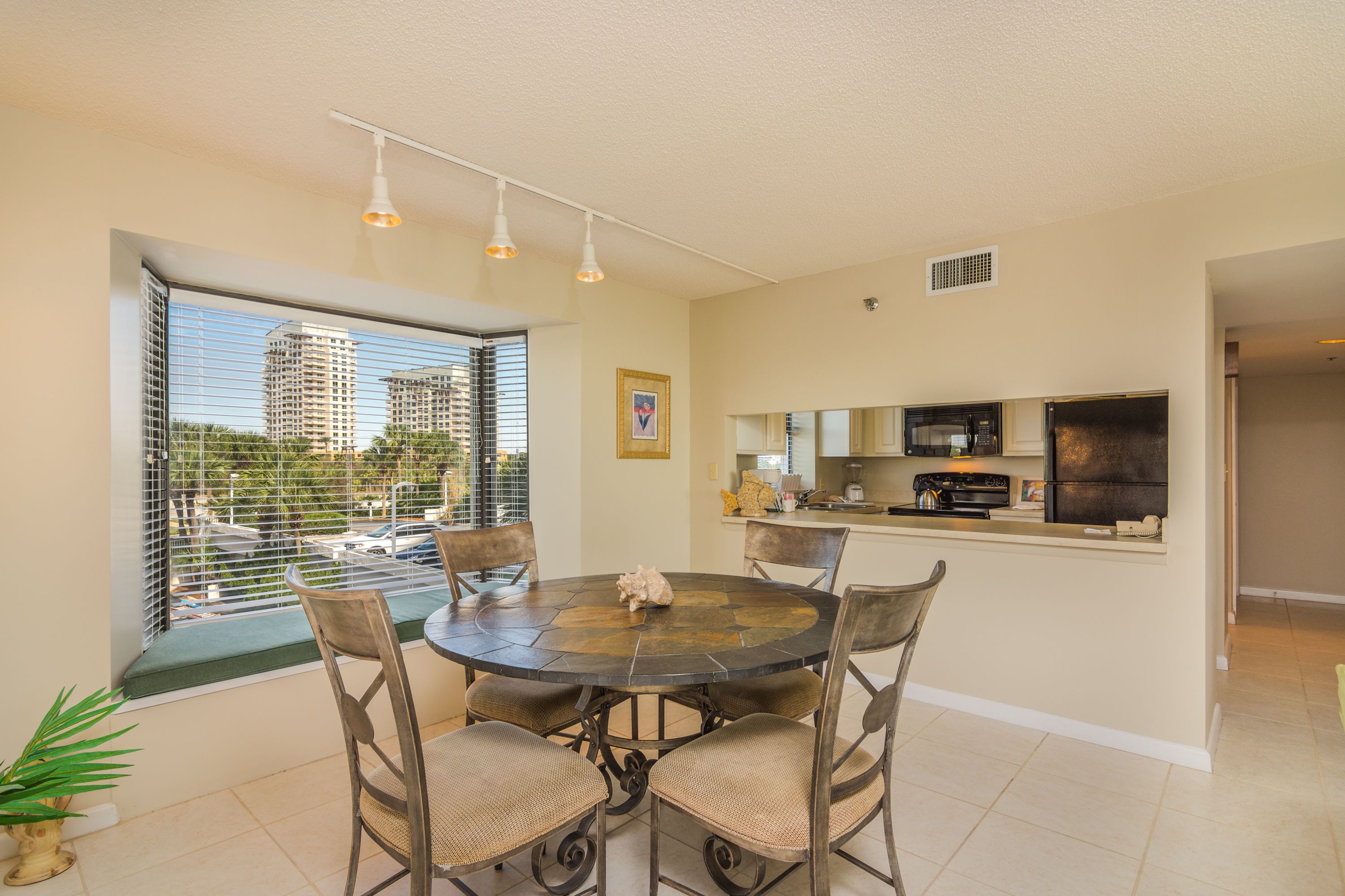 A dining area with a round table and chairs, a kitchen with appliances, and a view of tall buildings through a window.