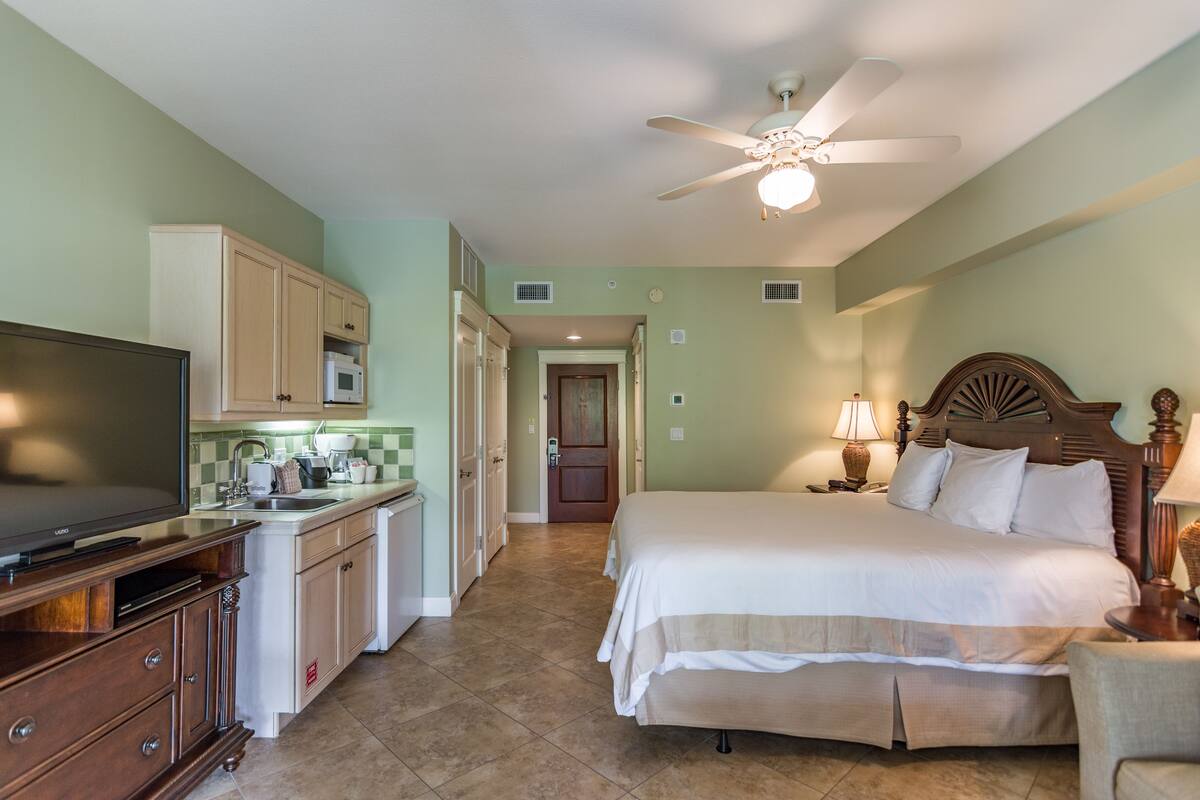 A hotel room with a bed, a TV, a kitchenette, and a ceiling fan.