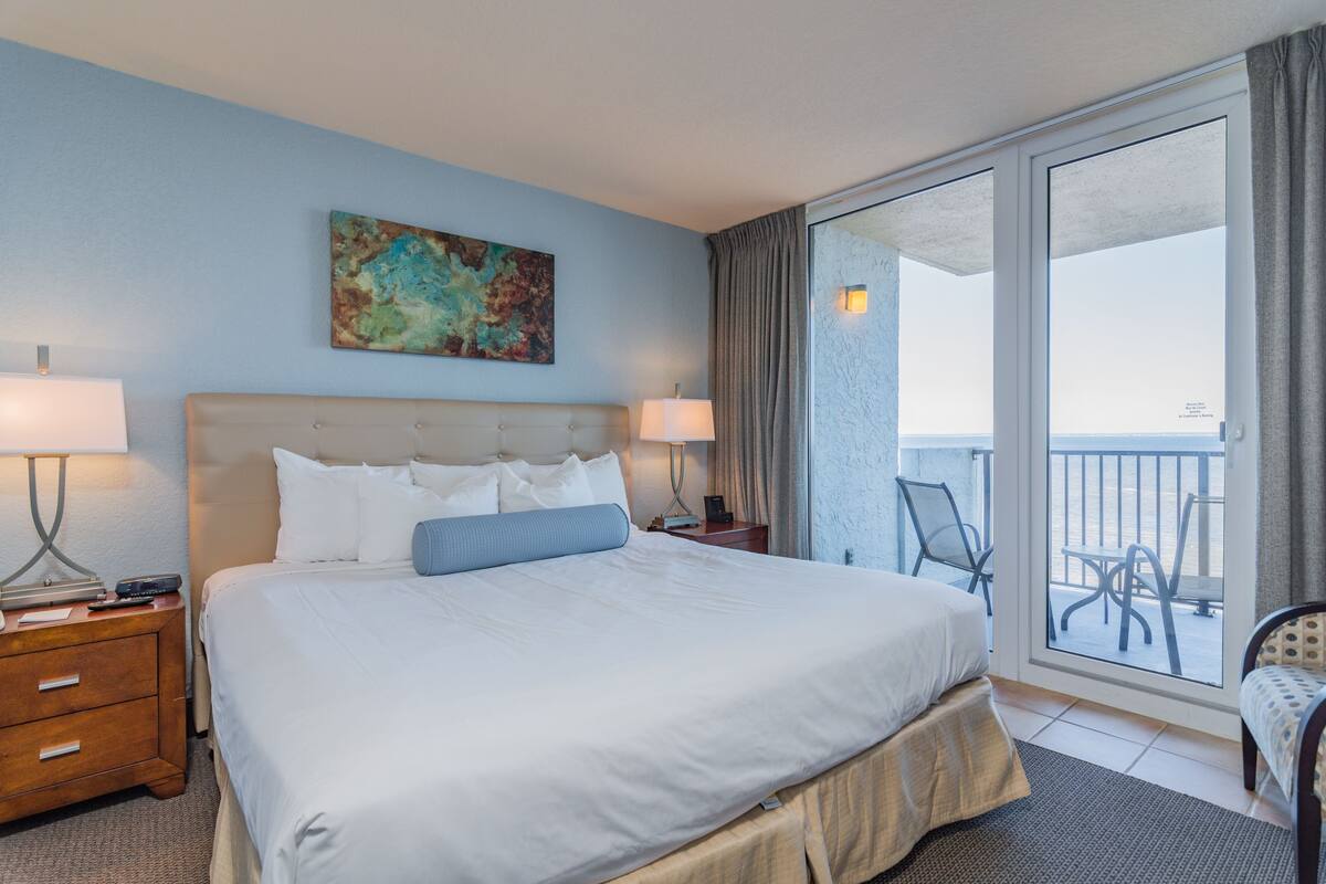 A hotel room with a large bed, two bedside tables with lamps, a painting on the wall, and a balcony with chairs.