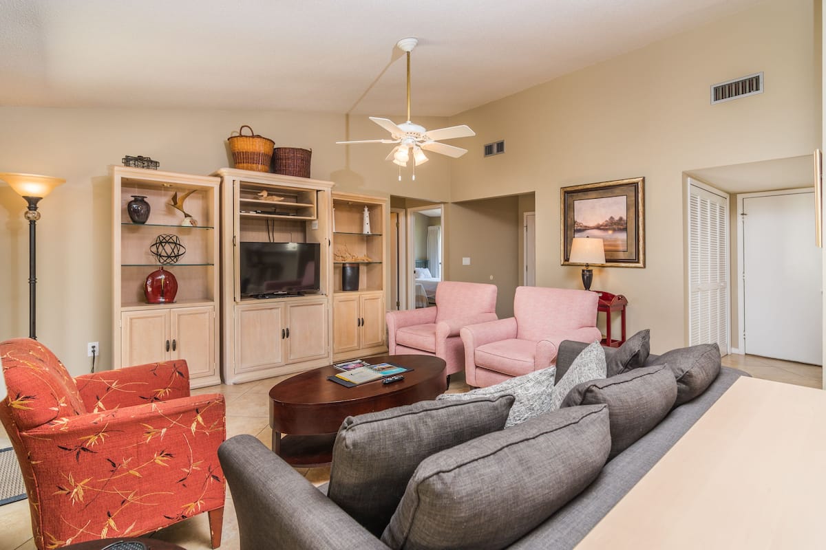 A living room with a sofa, armchairs, a coffee table, and a television. There is a ceiling fan, a lamp, and a framed picture on the wall.