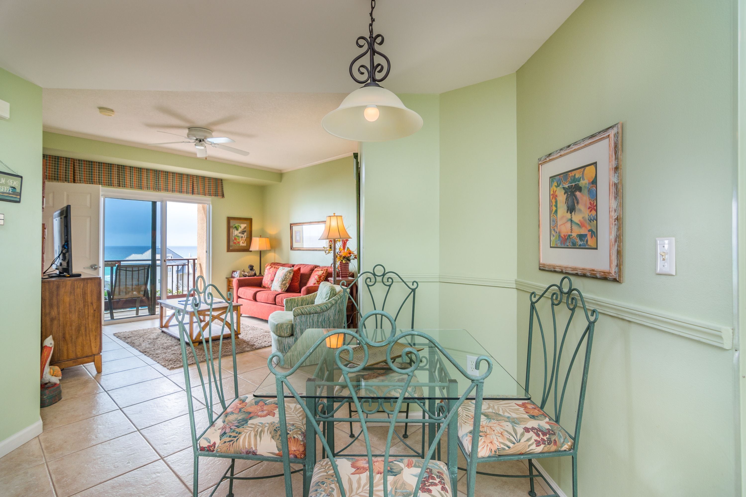 A dining area with a glass table and chairs, a ceiling fan, and a view of the ocean through a sliding door.