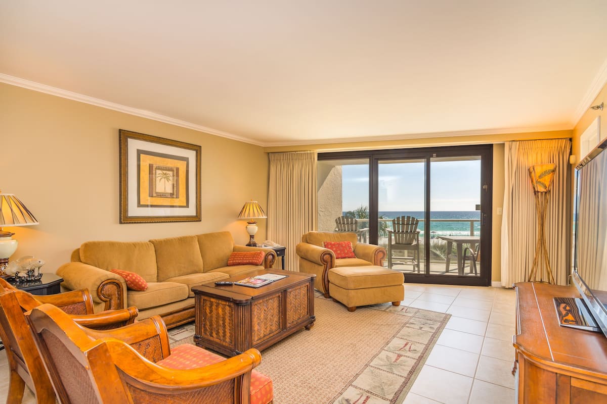 A living room with a sofa, armchairs, a coffee table, and a TV. There is a balcony with a view of the ocean.