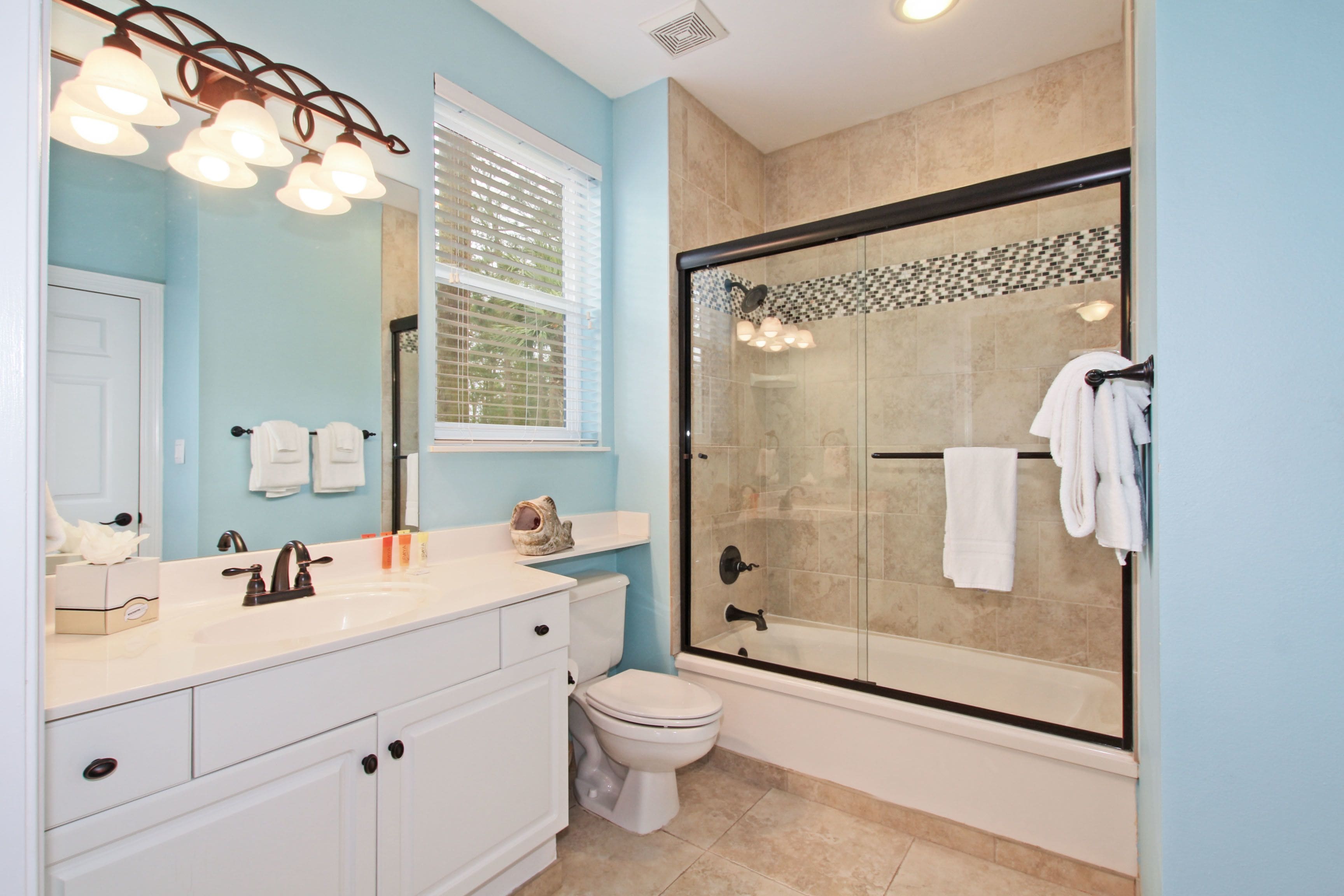 A bathroom with a white sink, mirror, and toilet, a glass-enclosed shower, and a window with blinds.