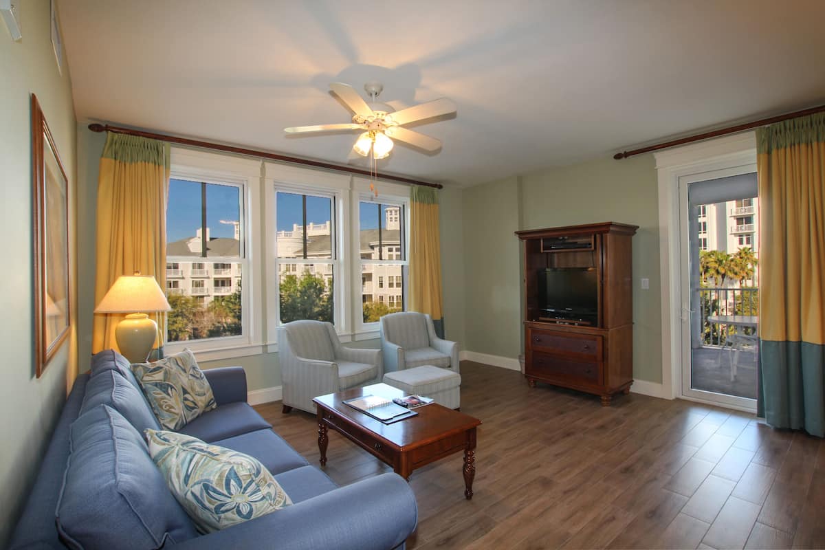 A living room with a sofa, chairs, a coffee table, a TV stand, and large windows with curtains.