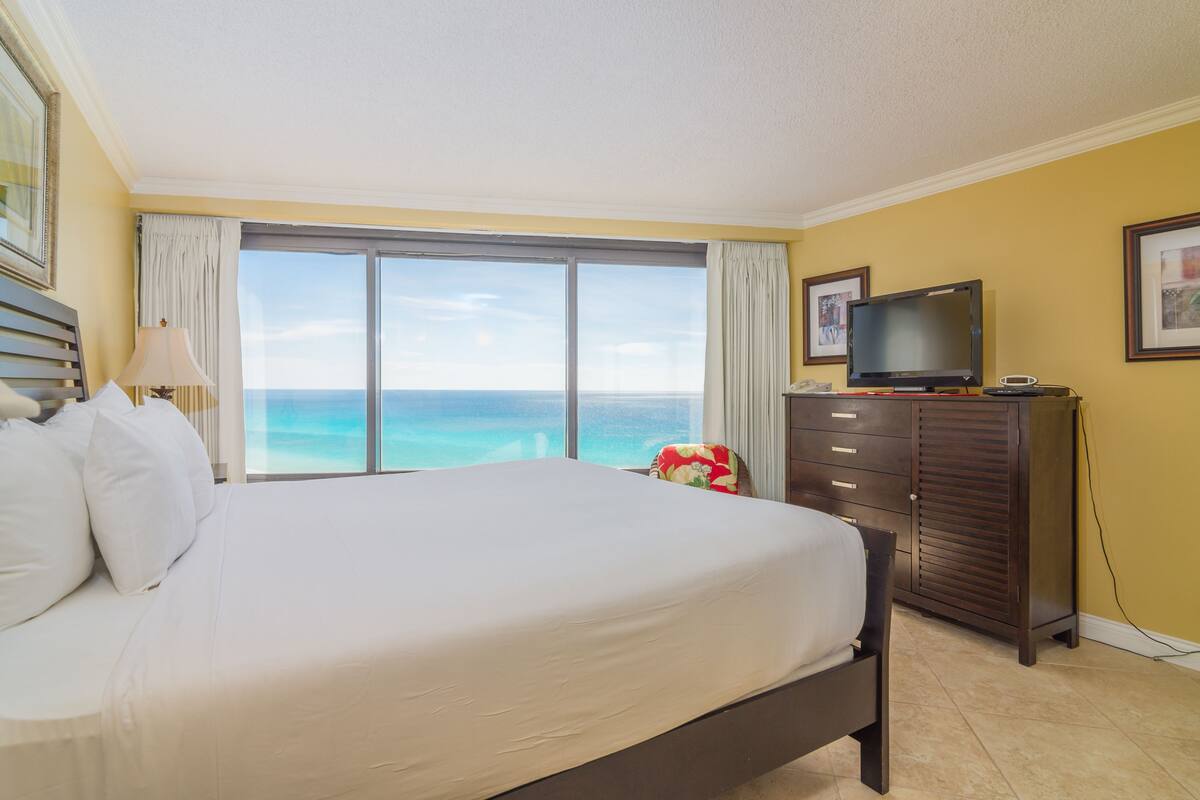 A hotel room with a large bed, a television, a dresser, and a view of the ocean.