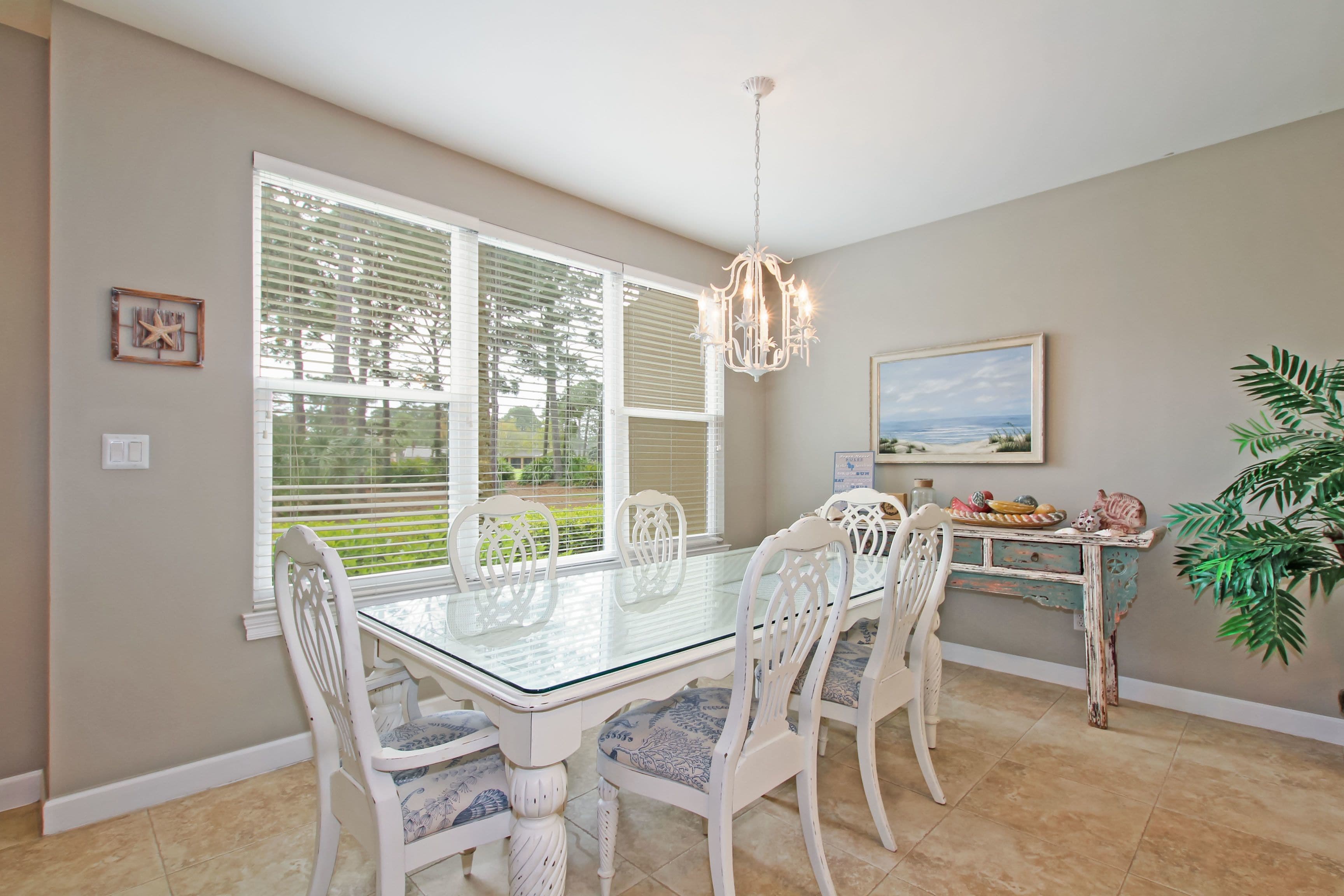 A dining area with a glass-top table, white chairs, a chandelier, a painting on the wall, and a plant.