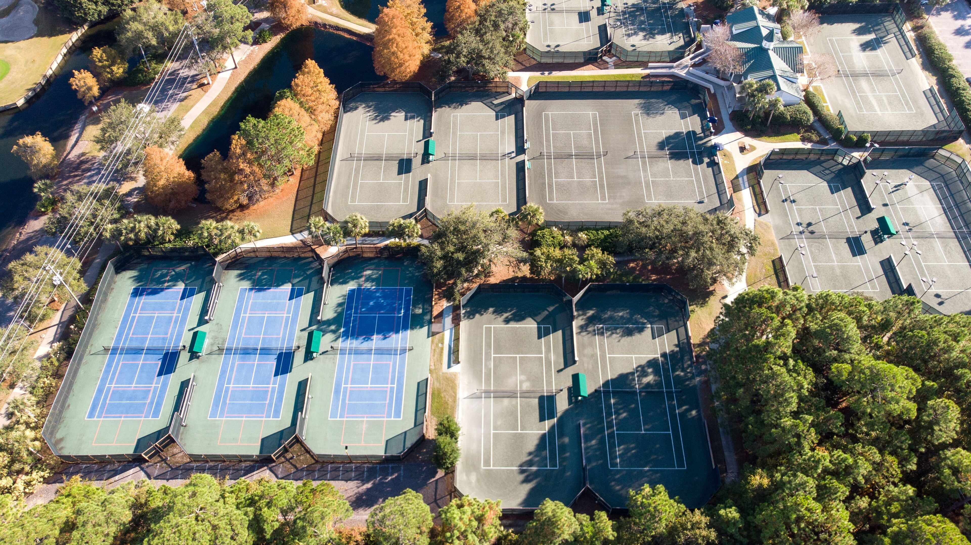 An aerial view of a sports complex with multiple tennis courts, surrounded by trees and a small body of water.
