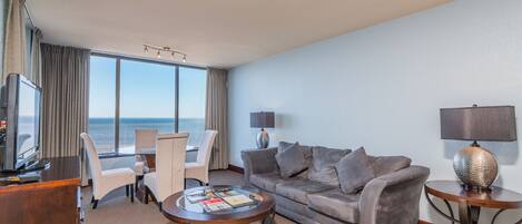 A hotel room with a sofa, armchairs, a coffee table, a TV, and a view of the sea.