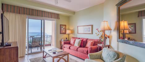 A living room with a sofa, armchair, coffee table, and a television. There is a balcony with a view of the ocean.