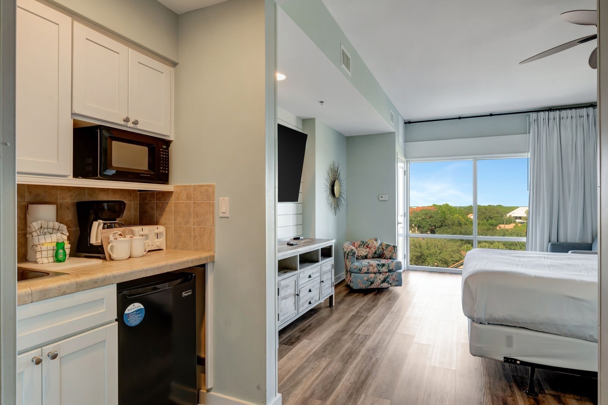 A modern kitchen with a dishwasher, microwave, and coffee maker, a dining area with a chair, and a bedroom with a bed and window.