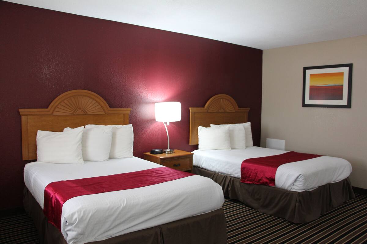 A hotel room with two beds, each with white linens and a red accent. There is a nightstand with a lamp between the beds, and a framed picture on the wall.