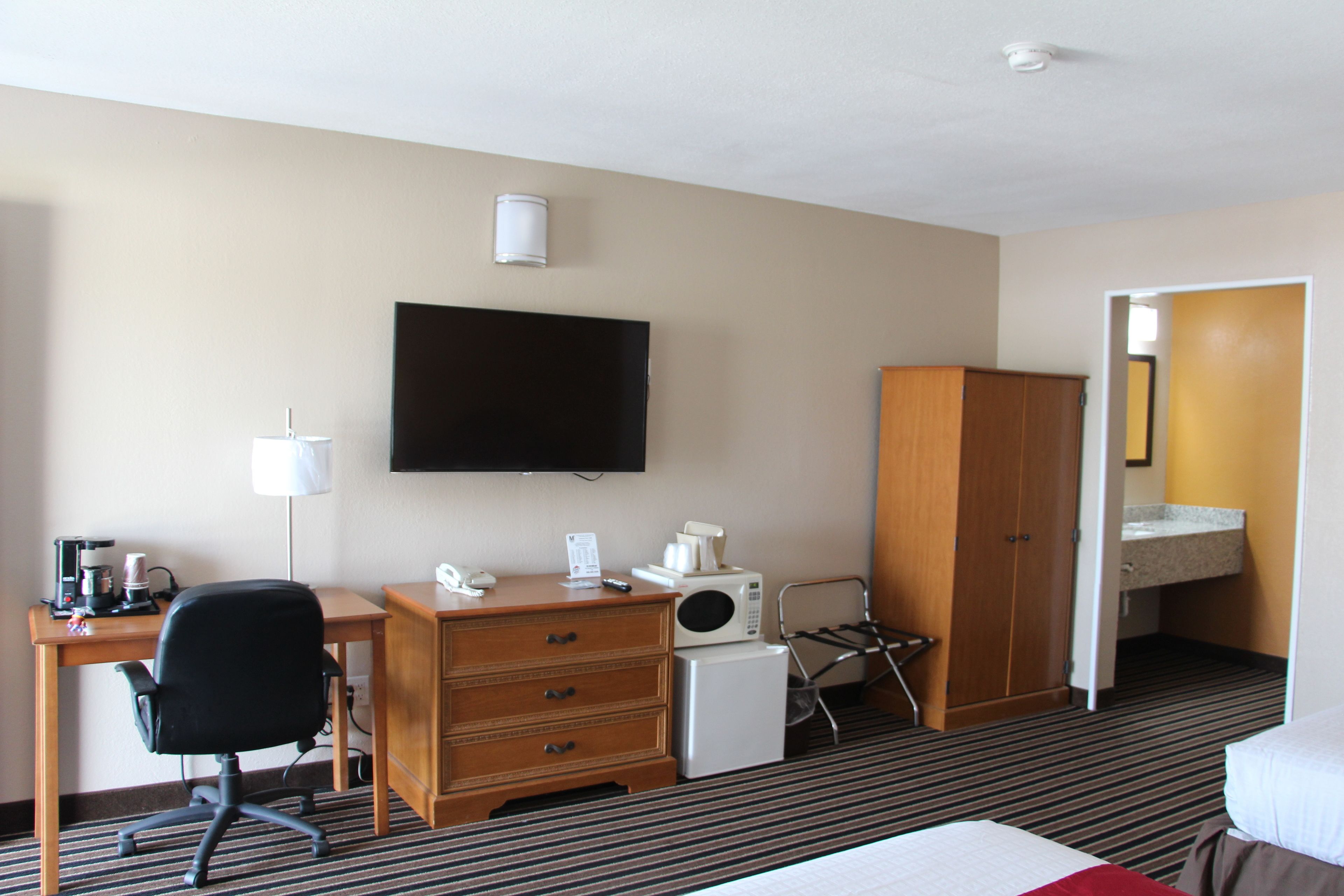 A hotel room with a bed, a desk with a chair, a television, a microwave, a refrigerator, a wardrobe, and a bathroom with a sink and mirror.