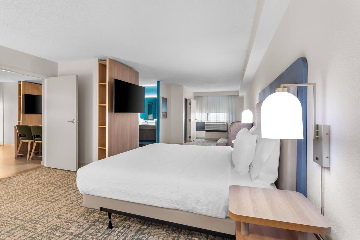 Et moderne hotelværelse med en stor seng, et skrivebord, et fladskærms-tv og et badeværelse, der kan anes gennem en åben dør.