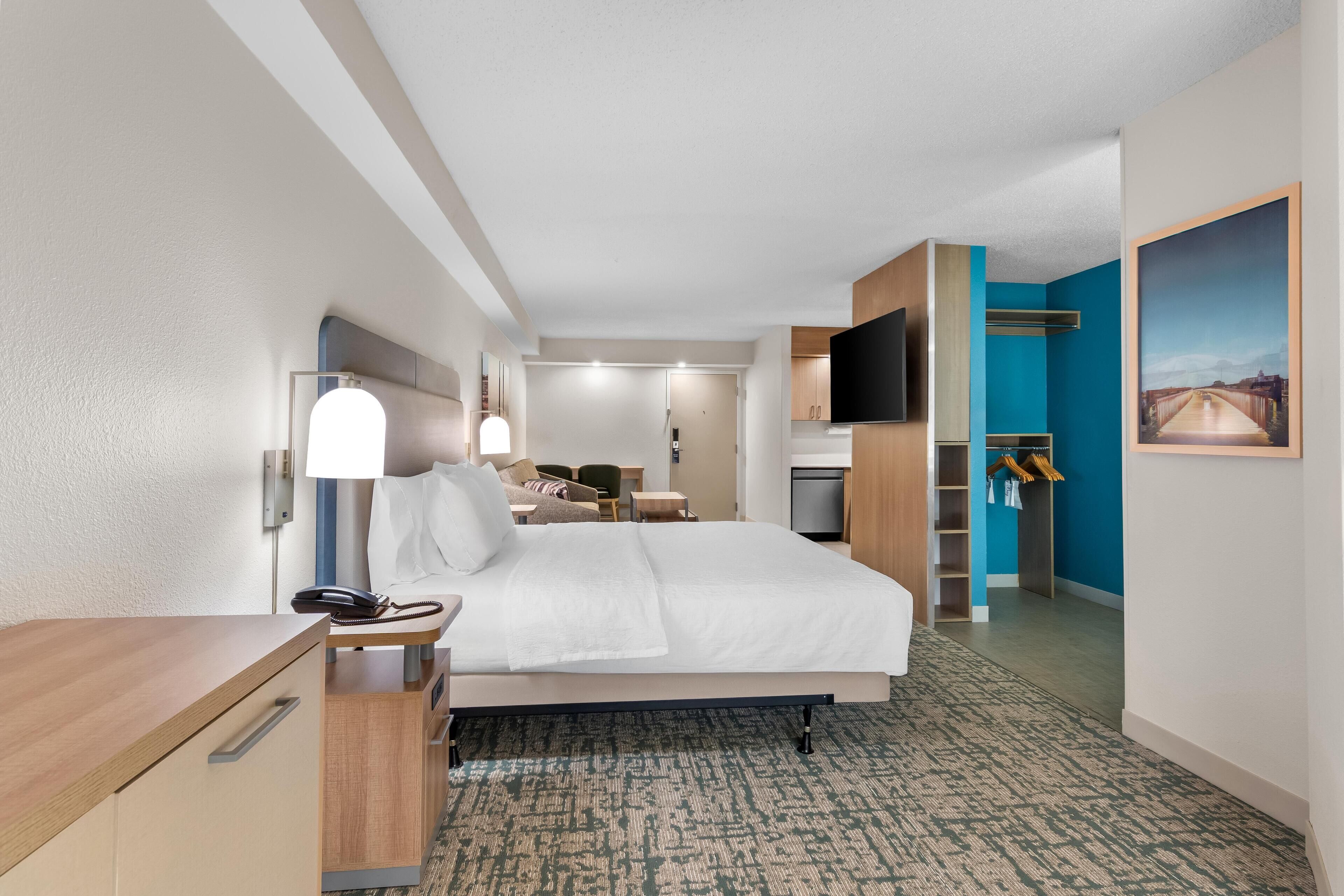 A hotel room with a bed, bedside table, lamp, and a TV mounted on the wall.