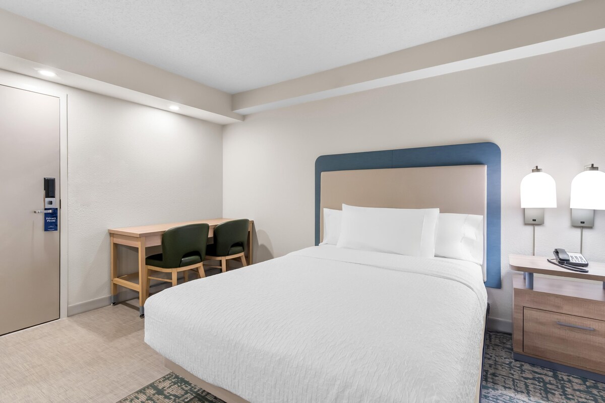 A hotel room with a bed, a desk, two chairs, a nightstand, and a phone.