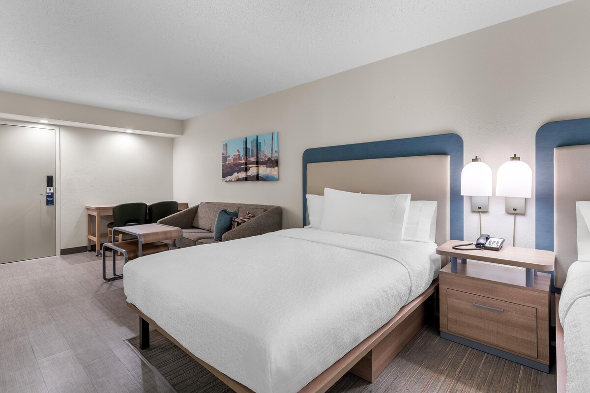 A hotel room with a bed, bedside tables, a desk, a chair, and a cityscape painting on the wall.