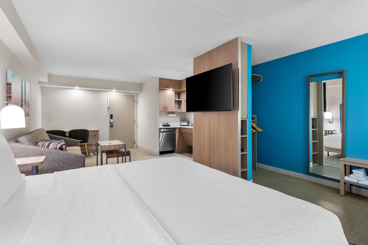 Et moderne hotelværelse med en stor seng, et fladskærms TV på væggen, et tekøkken og et badeværelse med et spejl.
