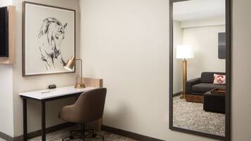 A hotel room with a desk, chair, and a large mirror reflecting a living area with a sofa and lamp.