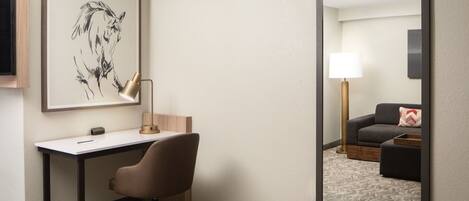 A hotel room with a desk, chair, and a large mirror reflecting a living area with a sofa and lamp.