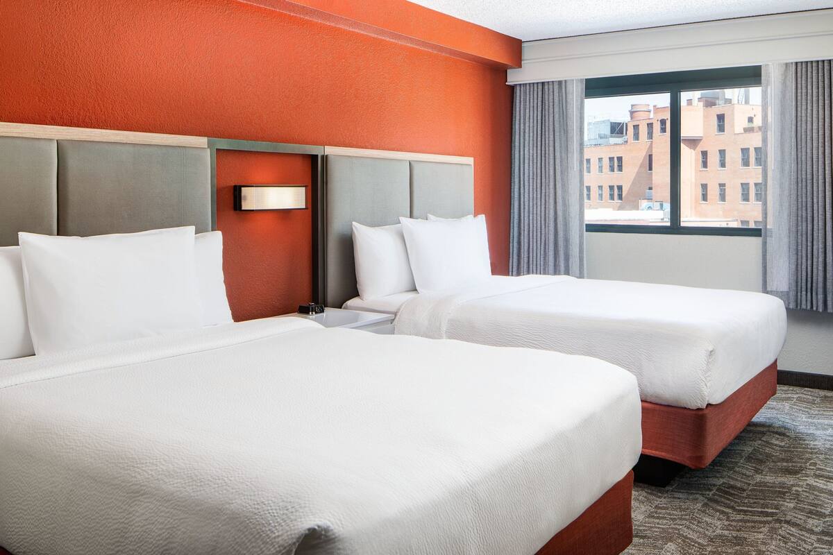A hotel room with two beds, a headboard, a nightstand, and a window with a view of buildings.