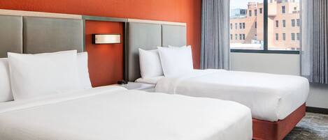 A hotel room with two beds, a headboard, a nightstand, and a window with a view of buildings.